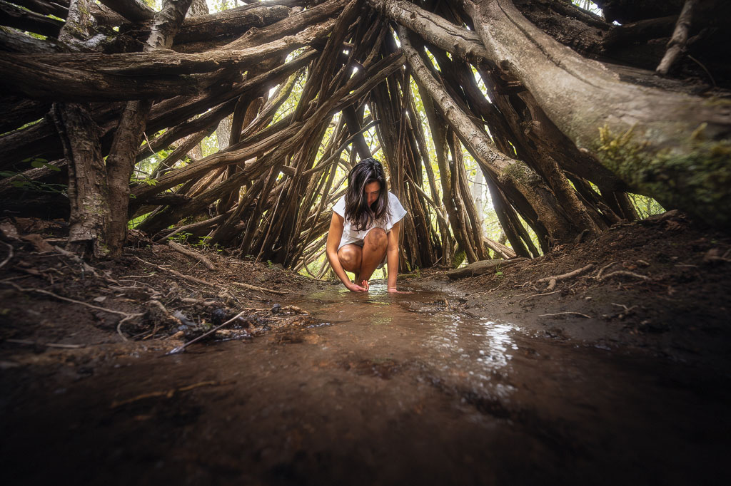 Femme interragissant avec l’eau dans un cours d’eau, sous un tipi fait de troncs d'arbre